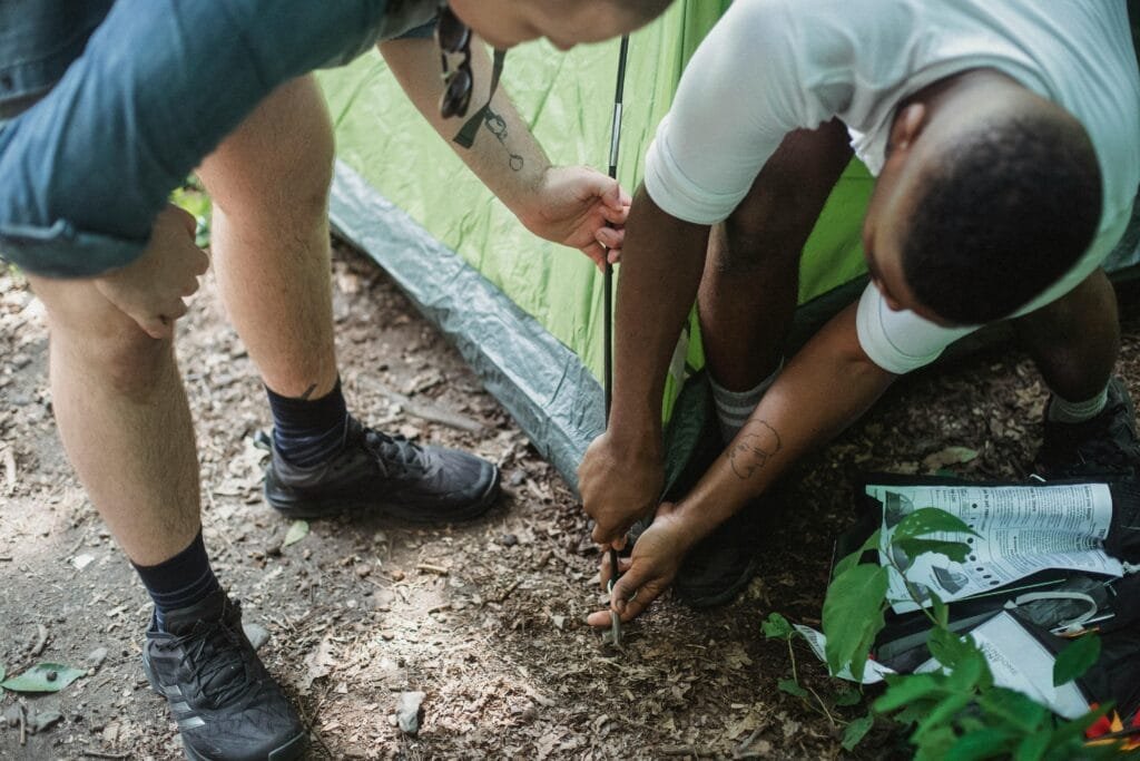 Two friends collaborate in setting up a camping tent in a lush forest setting, enjoying a summer adventure.