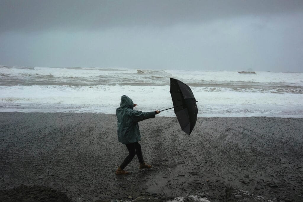 Full body of anonymous person in hood standing in wind with umbrella on sandy shore near stormy sea in rainy weather on a bad camping trip