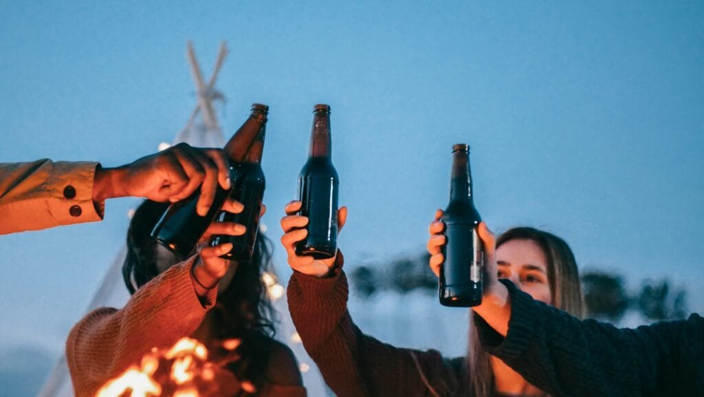 A group of friends celebrate with beer bottles around a campfire during sunset.